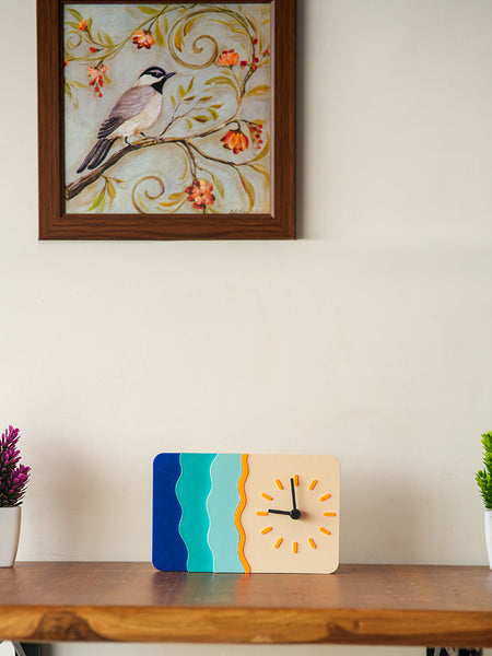 Decorative clock on a wooden shelf with two potted plants and a framed picture of a bird on a branch.