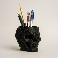 Black skull-shaped pen holder with various pens on a light beige background