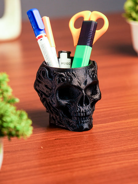 Black skull-shaped pen holder with stationery items on a wooden surface